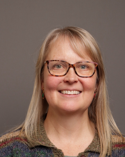 Head shot photo of Cindy Rosalbo with blonde hair and glasses, wearing a multi-colored sweater in front of a neutral background. 