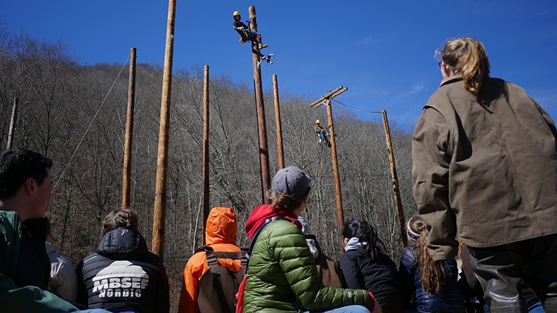 Students on the ground looking up at a power lineman climbing a utility pole